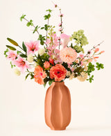In un vaso di terracotta si trova il colorato bouquet 'Yasmine's Choice' di fiori artificiali di seta, composto da gigli rosa, rose pesca e foglie verdi, realizzato con fiori di seta di alta qualità di Easyplants, tutti splendidamente disposti su uno sfondo chiaro.