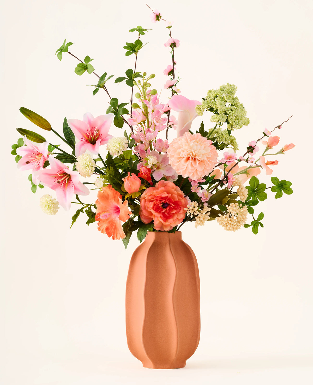 In un vaso di terracotta si trova il colorato bouquet 'Yasmine's Choice' di fiori artificiali di seta, composto da gigli rosa, rose pesca e foglie verdi, realizzato con fiori di seta di alta qualità di Easyplants, tutti splendidamente disposti su uno sfondo chiaro.