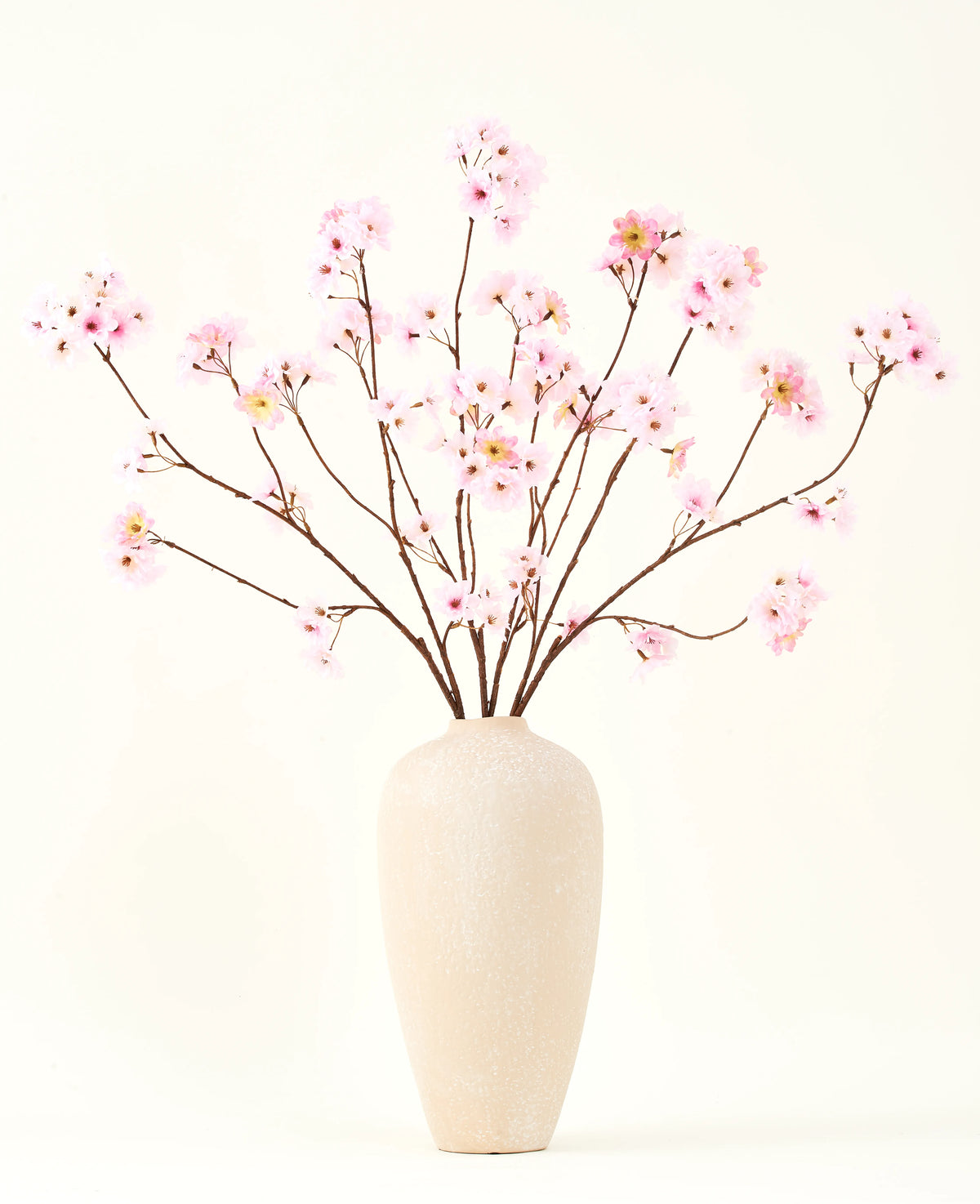 Il bouquet di fiori artificiali in seta Fiore di ciliegio rosa 114 cm ha rami lunghi e sottili con mazzi di fiori rosa chiaro in un vaso di ceramica beige, che formano un sorprendente contrasto con il semplice sfondo bianco.