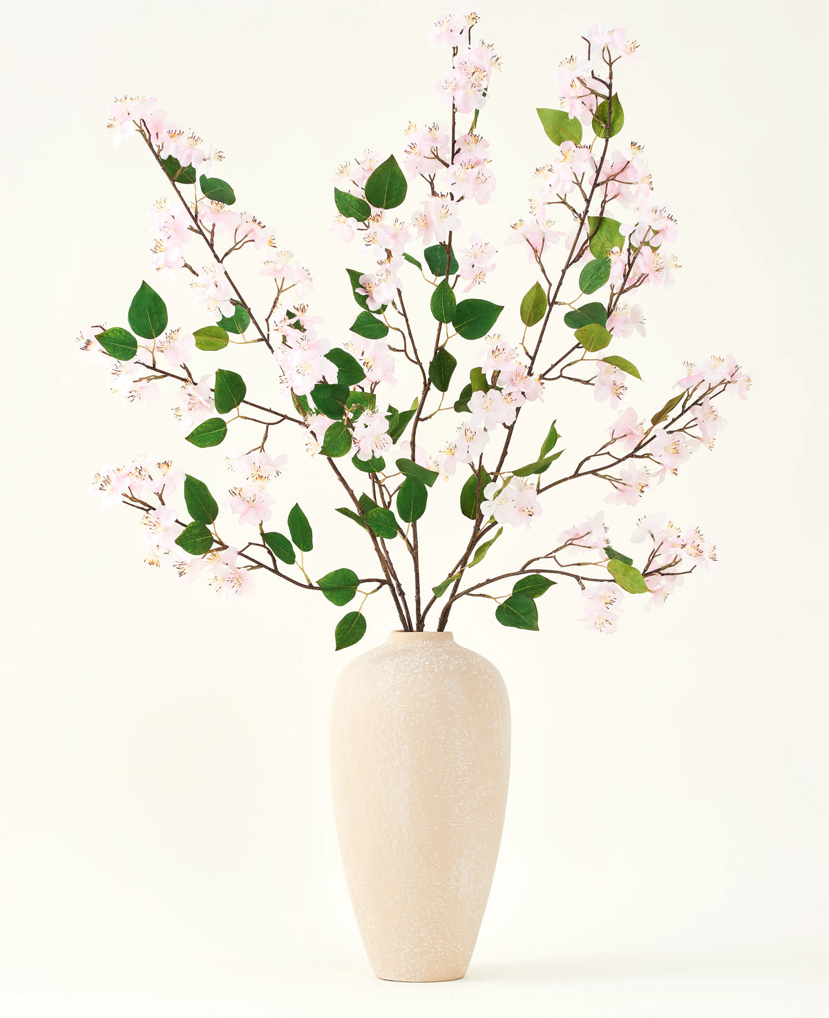 Un vaso in ceramica beige contiene diversi rami lunghi con il bouquet di fiori artificiali Silk di fiori di pesco rosa e foglie verdi, su uno sfondo bianco semplice, perfetto per un interno floreale artificiale.