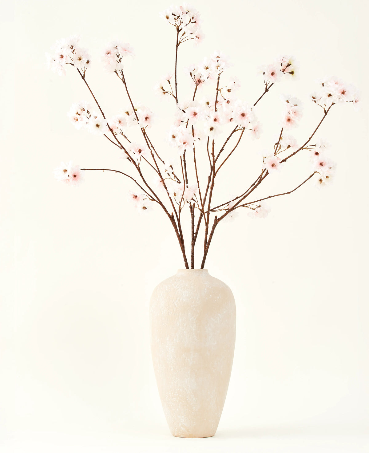 Il bouquet di fiori artificiali in seta Fiore di ciliegio blush 114 cm ha rami alti e delicati in un semplice vaso beige, che aggiunge un tocco morbido ed elegante ai tuoi interni su uno sfondo semplice e chiaro.