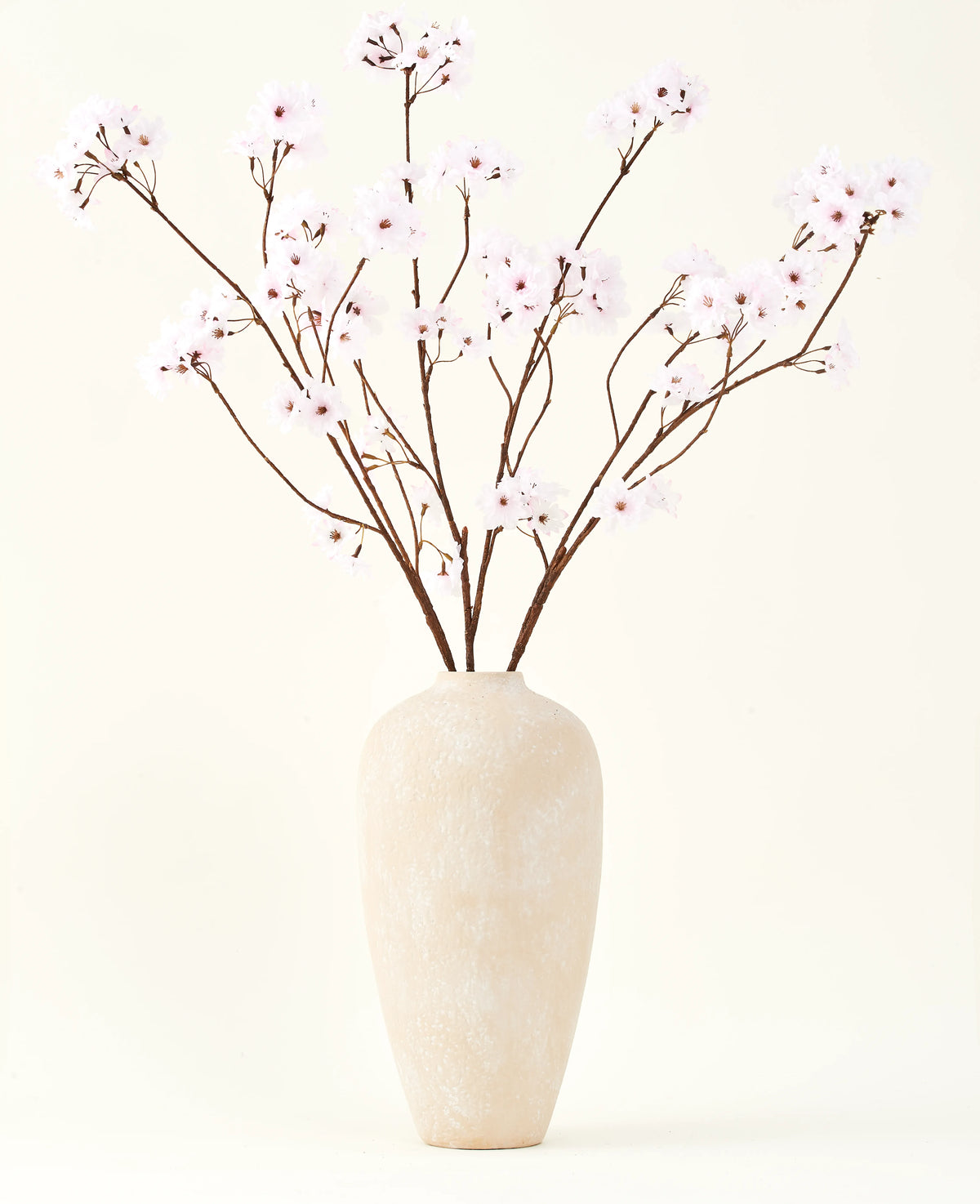 Il bouquet di fiori artificiali in seta Fiore di ciliegio rosa chiaro 114 cm si trova in un alto vaso di ceramica beige con rami lunghi e sottili e delicati fiori rosa chiaro, creando un sereno accento floreale artificiale su uno sfondo semplice.