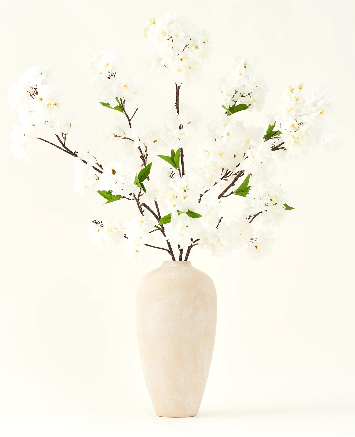 Il bouquet di fiori artificiali in seta Fiore di ciliegio crema 119 cm ha eleganti rami marroni con morbidi fiori finti bianchi e foglie verdi, esposti in un semplice vaso beige su uno sfondo chiaro.