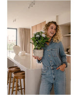 Una donna sorridente con una tuta di jeans si trova in una cucina moderna, con la pianta artificiale bianca Artificial Gardenia UV 39 cm tra le mani. Sgabelli di legno sono allineati lungo un piano di lavoro bianco e la luce del sole filtra attraverso una finestra sullo sfondo.