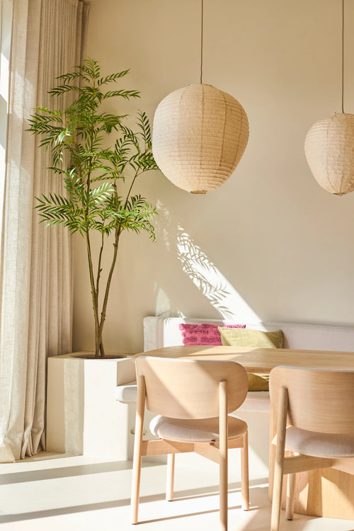 Uno spazio abitativo luminoso e minimalista con sedie in legno, un divano bianco con cuscini colorati, grandi finestre con tende, due lampade a sospensione lanterne di carta rotonde e la pianta artificiale Mahonia di 210 cm, che fornisce un tocco di verde. La luce del sole proietta ombre sui muri.