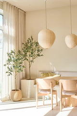 Una zona pranzo luminosa e minimalista è arredata con due sedie in legno chiaro, un tavolino, lanterne di carta e l'Ulivo Artificiale di 180 cm, che aggiunge un tocco di stile mediterraneo. Alberi interni in vaso, tende beige traslucide e luce solare che filtra dalle grandi finestre completano questo spazio rilassante.