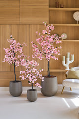 Tre piante in vaso del tipo Bougainvillea artificiale da 150 cm, di colore rosa con fiori rosa, sono disposte davanti a ripiani in legno, accanto a una moderna sedia bianca con cuscino e un candeliere decorativo.