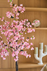 Una pianta di bouganville artificiale di 150 cm, di colore rosa e con fiori rosa, sta davanti a scaffali di legno su cui sono esposti un piatto decorativo e un moderno candeliere bianco con tre candele verdi.