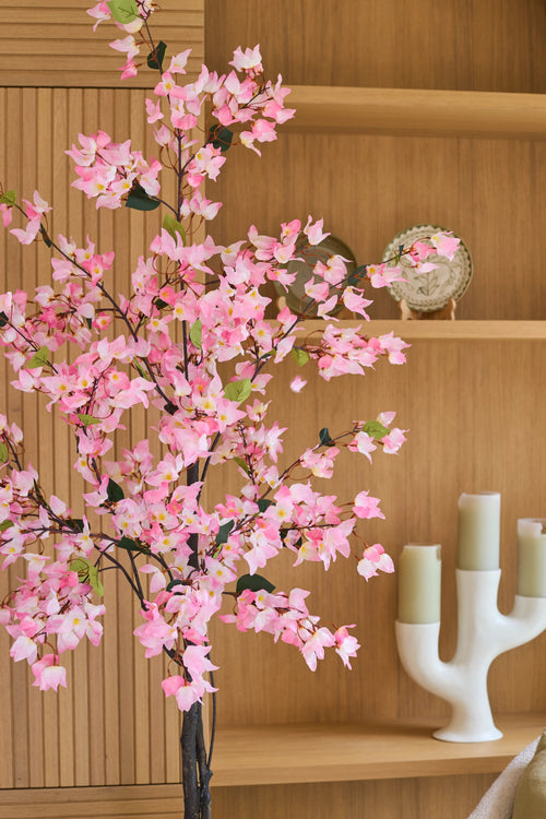Una pianta di bouganville artificiale di 150 cm, di colore rosa e con fiori rosa, sta davanti a scaffali di legno su cui sono esposti un piatto decorativo e un moderno candeliere bianco con tre candele verdi.