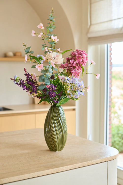 Su un'isola chiara della cucina, vicino a una finestra da cui entra la luce naturale, si trova un vaso verde con fiori rosa, viola e bianchi del bouquet di fiori artificiali in seta "Lavender Dreams".