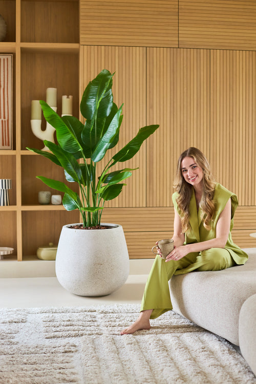 Una donna vestita di verde chiaro siede a piedi nudi su un divano, sorride e tiene in mano una tazza. Accanto a lei c'è la Pianta Artificiale Strelitzia Real Touch Deluxe 150 cm, mentre assi di legno e oggetti decorativi donano allo sfondo un aspetto caldo.