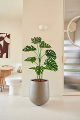 La pianta artificiale Monstera da 120 cm è collocata in uno spazio abitativo moderno e minimalista con pareti neutre, uno specchio rotondo, mobili in legno e quadri astratti.
