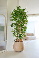 La pianta artificiale di bambù 210 cm si trova in un soggiorno luminoso e moderno con un divano beige, tende bianche e un arredamento minimalista.