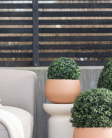 Tre piante artificiali topiarie rotonde di bosso stile romano UV D30 cm si trovano su un patio vicino a una sedia da esterno beige, contro una moderna recinzione a doghe nere e alberi spogli.