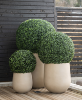 Tre arbusti artificiali di bosso artificiale in stile romano UV D40 cm in grandi vasi beige si trovano su una terrazza in legno, contro una staccionata in legno scuro con doghe orizzontali.
