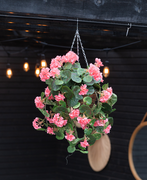 La Begonia artificiale rosa in cesto UV 70 cm, con foglie verdi lussureggianti e fiori rosa, è appesa a una catena. Calde luci radenti e uno specchio rotondo accentuano lo sfondo in legno scuro.