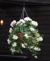 La Begonia artificiale bianca in cesto UV 70 cm, con foglie verdi lussureggianti e fiori bianchi, è appesa a una catena di metallo davanti a una parete di legno scuro con una lampada luminosa sullo sfondo.