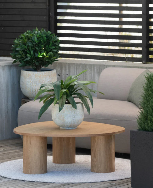 Su un piccolo tappeto accanto a un divano grigio in una terrazza c'è un tavolino in legno con sopra una pianta artificiale Bird's Deluxe di 60 cm, con alcune piante in vaso sullo sfondo.