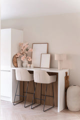 Un bar minimalista con due sgabelli beige con cuscini, un bancone bianco, opere d'arte incorniciate, una lampada da tavolo, decorazioni dai toni neutri e vasi di fiori di ciliegio artificiali da 100 cm rosa chiaro contro una parete di colore chiaro.