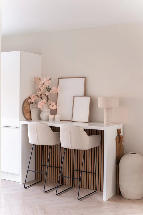 Un bar minimalista con due sgabelli beige con cuscini, un bancone bianco, opere d'arte incorniciate, una lampada da tavolo, decorazioni dai toni neutri e vasi di fiori di ciliegio artificiali da 100 cm rosa chiaro contro una parete di colore chiaro.