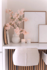 Una scrivania minimale con una sedia bianca, due candele, vassoi decorativi, arte astratta incorniciata e un fiore di ciliegio artificiale di 100 cm rosa chiaro in un vaso bianco su una superficie bianca.
