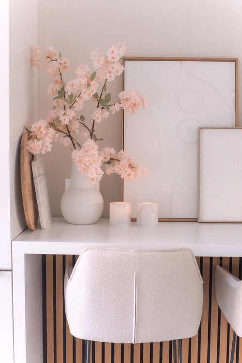 Una scrivania minimale con una sedia bianca, due candele, vassoi decorativi, arte astratta incorniciata e un fiore di ciliegio artificiale di 100 cm rosa chiaro in un vaso bianco su una superficie bianca.