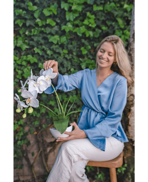 Una donna sorridente con una camicetta azzurra e pantaloni bianchi si siede su uno sgabello, tiene l'orchidea artificiale bianca di 50 cm in un vaso e tocca delicatamente i fiori. Si siede su uno sfondo di fogliame verde.
