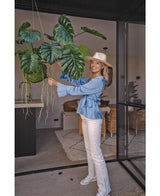 All'interno c'è una donna sorridente con cappello, camicetta blu e pantaloni bianchi che tiene in mano una pianta pensile artificiale Monstera Deluxe da 60 cm con grandi foglie verdi, circondata da un arredamento moderno per un'atmosfera tropicale con piante decorative.