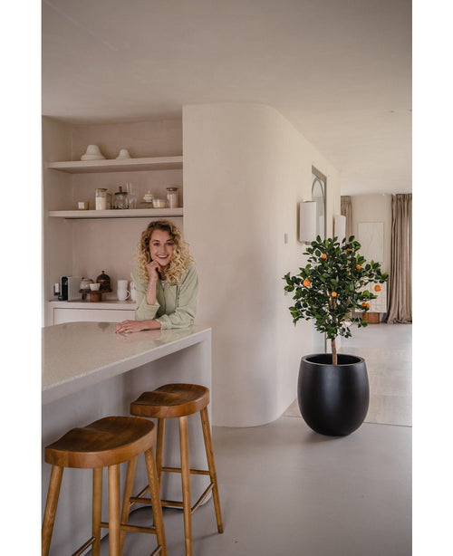 Una donna dai capelli biondi e ricci sorride, appoggiata al bancone. Accanto a lei, due sgabelli in legno e un albero di arancio artificiale di 120 cm donano un'atmosfera fresca alla cucina moderna e luminosa con ripiani e piatti dietro di essa.
