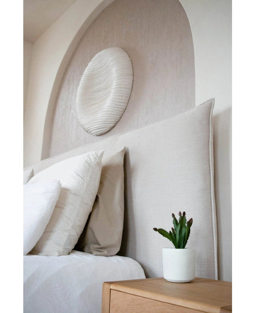 Una camera da letto moderna ha una testiera imbottita beige, cuscini bianchi, una pianta grassa artificiale di 20 cm su un comodino in legno e decorazioni murali rotonde strutturate sopra il letto.