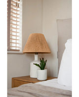 Un comodino con una lampada bianca con paralume intrecciato beige e una pianta grassa artificiale di 20 cm si trova accanto a un letto con lenzuola bianche e beige, vicino a una finestra con persiane in legno.
