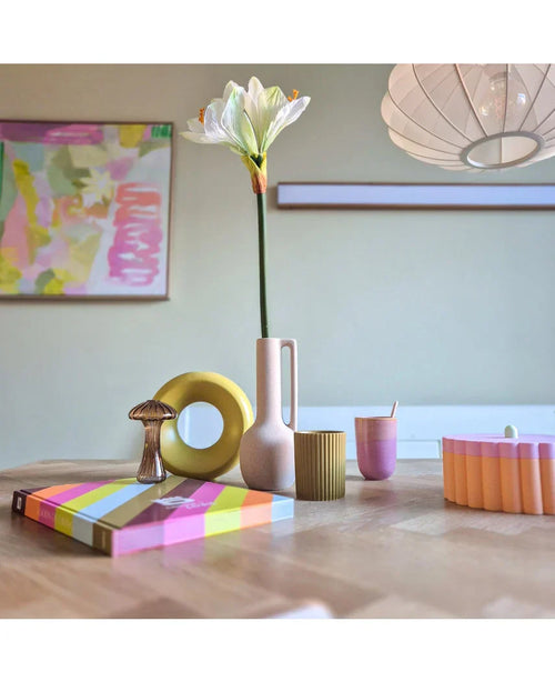 Un moderno tavolo in legno mostra oggetti colorati, tra cui un vaso rosa contenente il fiore artificiale Amaryllis Real Touch bianco da 66 cm, una scultura ad anello gialla, libri a strisce e una tazza rosa. L'arte astratta accentua la parete di fondo.