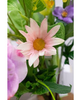 Un primo piano del fiore artificiale Aster di 60 cm con petali rosa con un centro giallo, circondato da foglie verdi e altri fiori, incluso uno viola, sullo sfondo.