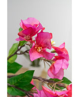 Primo piano del fiore artificiale Bougainvillea 90 cm viola: vivaci fiori artificiali di bouganville viola con foglie verdi su un ramo, su uno sfondo morbido e chiaro.