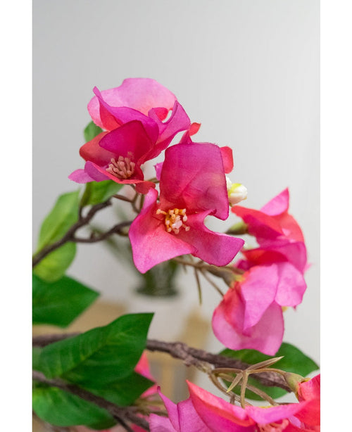 Primo piano del fiore artificiale Bougainvillea 90 cm viola: vivaci fiori artificiali di bouganville viola con foglie verdi su un ramo, su uno sfondo morbido e chiaro.