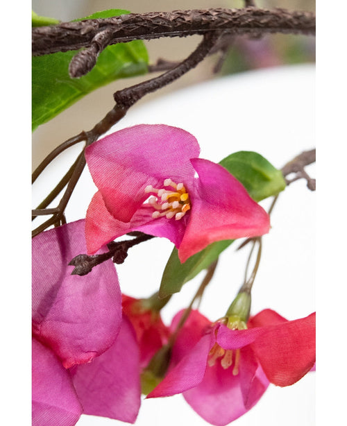 Primo piano del fiore artificiale Bouganvillea 90 cm viola, con vivaci fiori artificiali viola con stami gialli e foglie verdi su un ramo marrone testurizzato, su uno sfondo bianco sfocato.