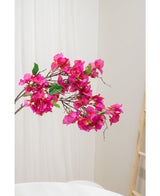 Il fiore artificiale Bougainvillea viola da 90 cm, con vivaci fiori viola e foglie verdi, è mostrato su uno sfondo neutro chiaro accanto a una scala di legno sulla destra.