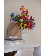 Un mazzo di fiori di garofano artificiale di 70 cm di colore blu in un vaso di ceramica bianca si trova su una scrivania bianca accanto a libri impilati e una piccola ciotola decorativa, con un vassoio rotondo di legno appoggiato al muro dietro di esso.