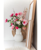 Un fiore artificiale margherita bianco/rosa da 60 cm si trova in un vaso bianco su una superficie bianca accanto all'arte in una cornice di legno, con un vassoio rotondo di legno visibile sullo sfondo.