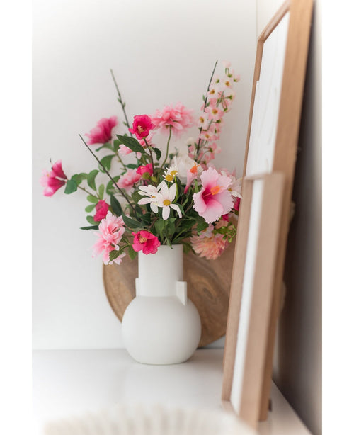 Un fiore artificiale margherita bianco/rosa da 60 cm si trova in un vaso bianco su una superficie bianca accanto all'arte in una cornice di legno, con un vassoio rotondo di legno visibile sullo sfondo.