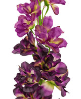 Un primo piano del fiore artificiale Delphinium Purple da 79 cm, un alto stelo artificiale con molteplici fiori viola e viola scuro con un cuore verde e giallo, su sfondo bianco.