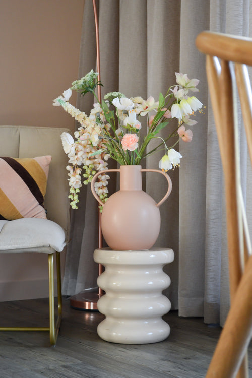 Il fiore artificiale Fritillary bianco da 94 cm si trova in un vaso di ceramica rosa chiaro con manici, posizionato su una base bianca ondulata accanto a una sedia moderna con cuscino a righe e tende neutre in una stanza elegante.