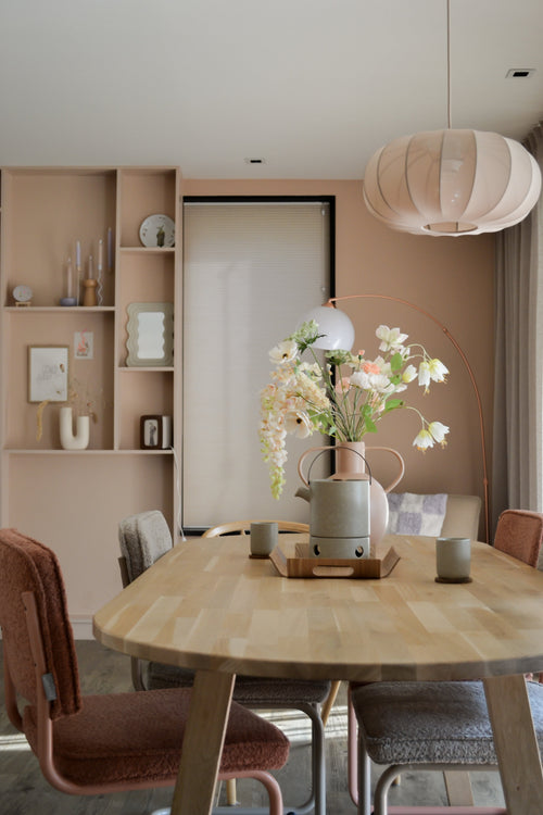 Una sala da pranzo moderna con un tavolo in legno chiaro, quattro sedie con cuscini, il fiore artificiale Fritillary bianco da 94 cm in un vaso, tazze in ceramica, pareti rosa pastello, mensole integrate con decorazioni e una grande lampada a sospensione rotonda sopra la testa.