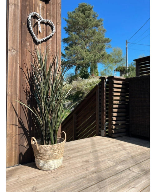 L'erba artificiale Bordeaux UV 120 cm è posizionata in un cestino intrecciato su una terrazza di legno vicino a una parete di legno con una ghirlanda a forma di cuore, con alberi e un cielo azzurro sullo sfondo.