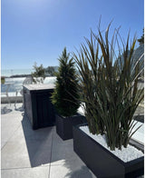 Erba artificiale Bordeaux UV 120 cm in fioriere rettangolari con pietre bianche decora una moderna terrazza all'aperto su uno sfondo di cielo azzurro e alberi in lontananza.