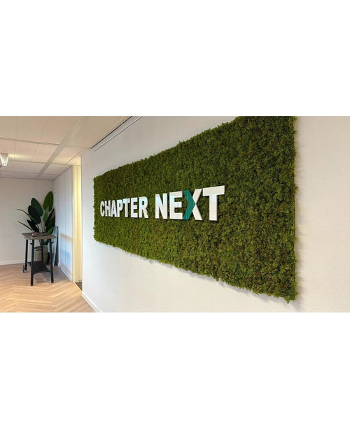 Un lussuoso accento interno: un espositore da parete verde con cornici UV 50x50 cm Kunsthaag Nordic Moss, con "CAPITOLO SUCCESSIVO" in grassetto lettere 3D. Un tavolo decorato con piante completa la raffinata scena.