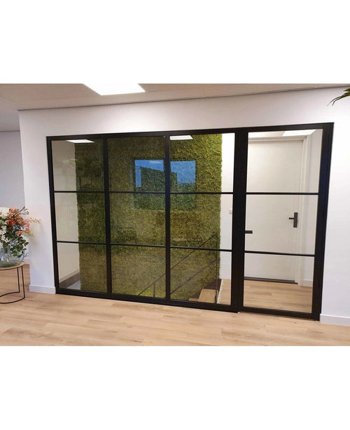 Un interno moderno e di lusso con una parete di vetro nero rivela una parete verde di Muschio Nordico dell'Aia Artificiale 50x50 cm UV, una scala dietro di essa, pavimenti in legno chiaro e un'elegante composizione floreale sulla sinistra.
