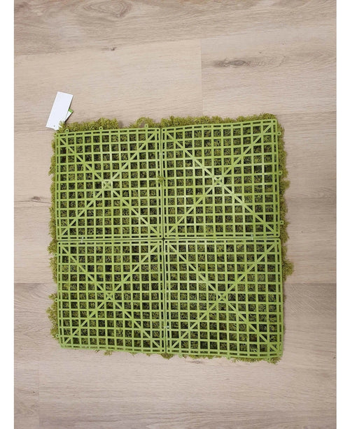Il muschio artificiale Haag Nordic Moss 50x50 cm UV è un tappeto in erba artificiale verde con una griglia posteriore in plastica visibile, ideale per interni lussuosi o per creare una parete verde. Poggia su un pavimento di legno chiaro con una piccola etichetta bianca in un angolo.