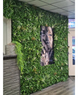 Una parete rivestita con siepe artificiale Savanne 100x100 cm, con verde protetto dai raggi UV in fioriera in plastica Ø100, ha al centro un grande ritratto in bianco e nero. Gli interni moderni hanno un soffitto piastrellato e un bancone in legno sulla sinistra.