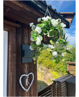 La Begonia artificiale bianca in cesto UV 70 cm è appesa a una parete di legno, con un cuore bianco decorativo sotto. Il verde, una terrazza in legno e un cielo azzurro parzialmente nuvoloso completano lo sfondo.